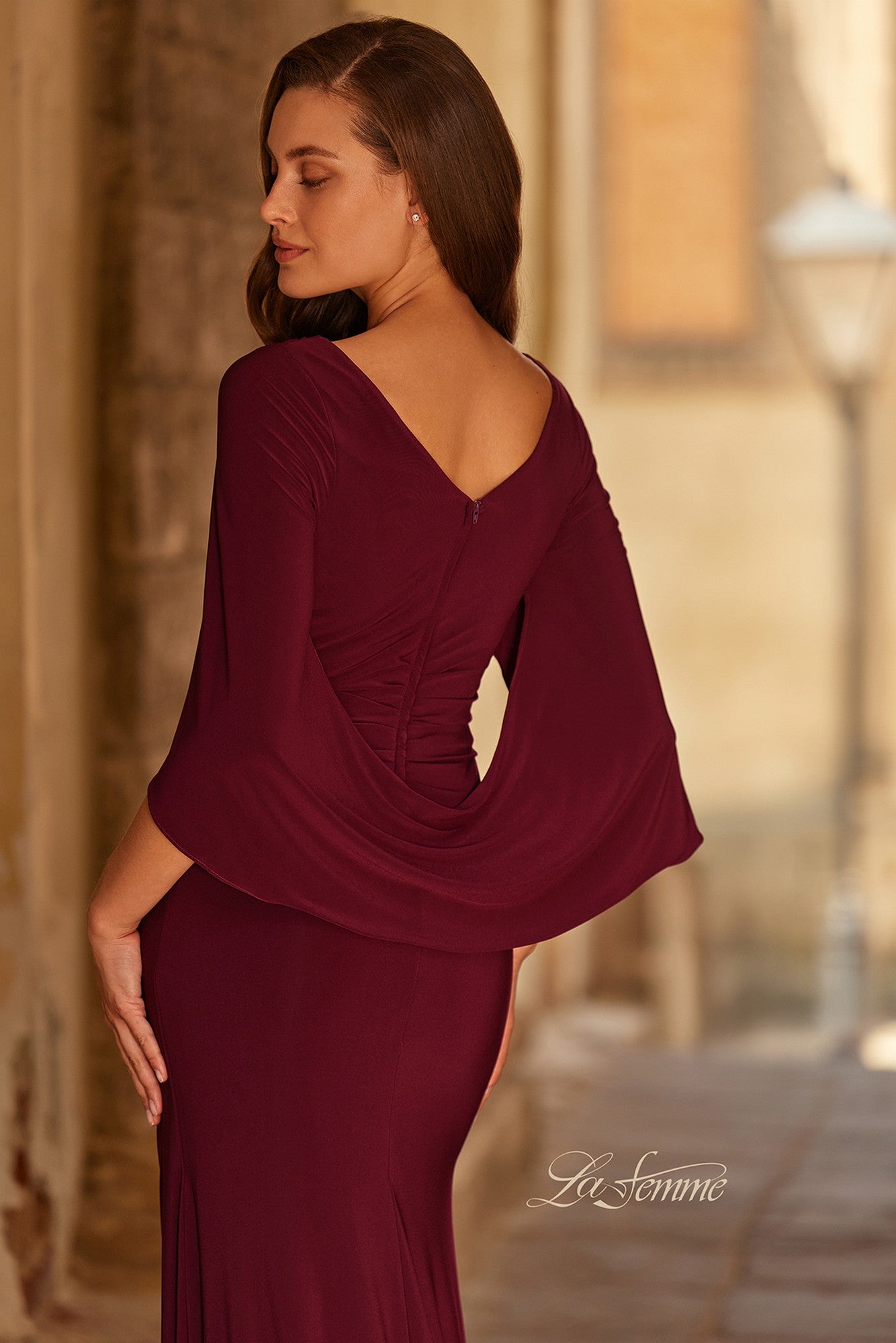 A woman wears the Long Formal Dress: La Femme 33148, a burgundy jersey evening gown with draped cape-style sleeves. She stands sideways outdoors, gazing downward.